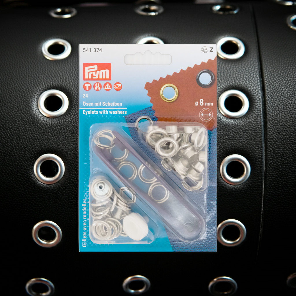Prym Ösen mit Scheiben, Ø 11 mm silberfarbig und mit Werkzeug in durchsichtiger Verpackung auf schwarzem Lederhintergrund mit kleinen Ösen