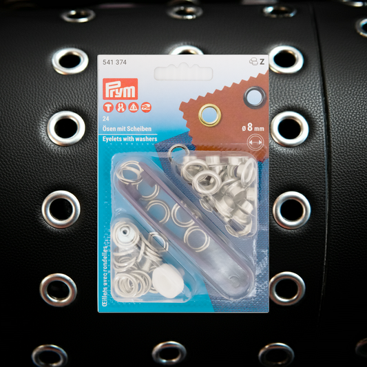 Prym Ösen mit Scheiben, Ø 11 mm silberfarbig und mit Werkzeug in durchsichtiger Verpackung auf schwarzem Lederhintergrund mit kleinen Ösen