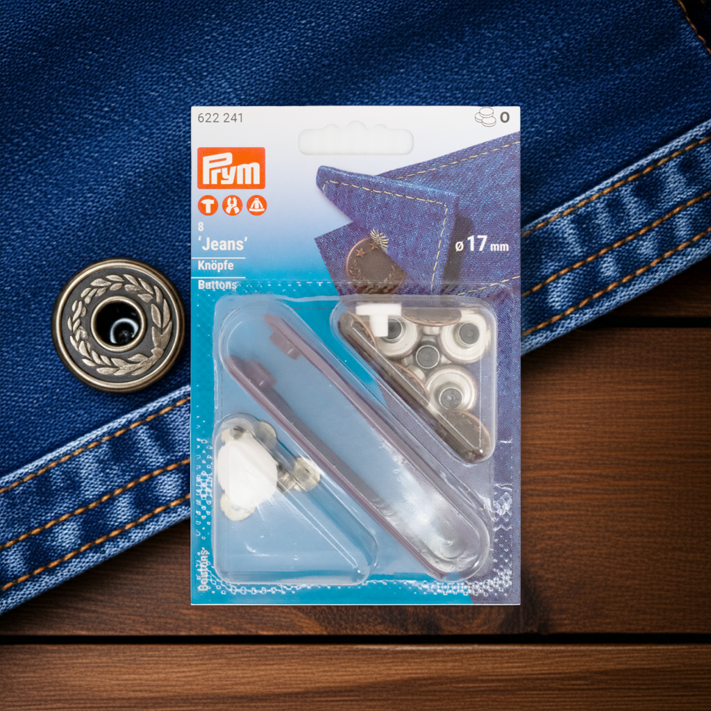 CasaBellissimo Prym Jeansknöpfe "Lorbeerkranz", nähfrei, Ø 17 mm in silberfarbig und mit Werkzeug in der Blister Verpackung auf Holz Untergrund und im Hintergrund eine Jeans mit einem Lorbeerkranz Knopf